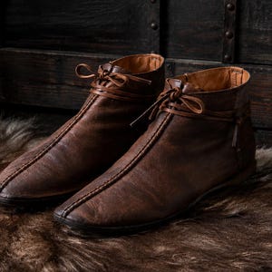 May include: A pair of dark brown leather ankle boots featuring a center seam running from the toe to the collar, a rounded toe shape, and thin leather laces tied in a bow at the top of the shaft. The interior lining is a lighter tan color.
