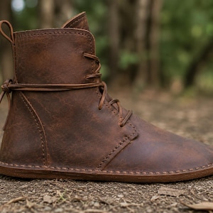 May include: A brown leather ankle boot featuring a rounded toe, a flat sole, and a lace-up closure that wraps around the ankle. The boot has visible stitching along the seams and a small pull tab at the top of the heel for ease of use.