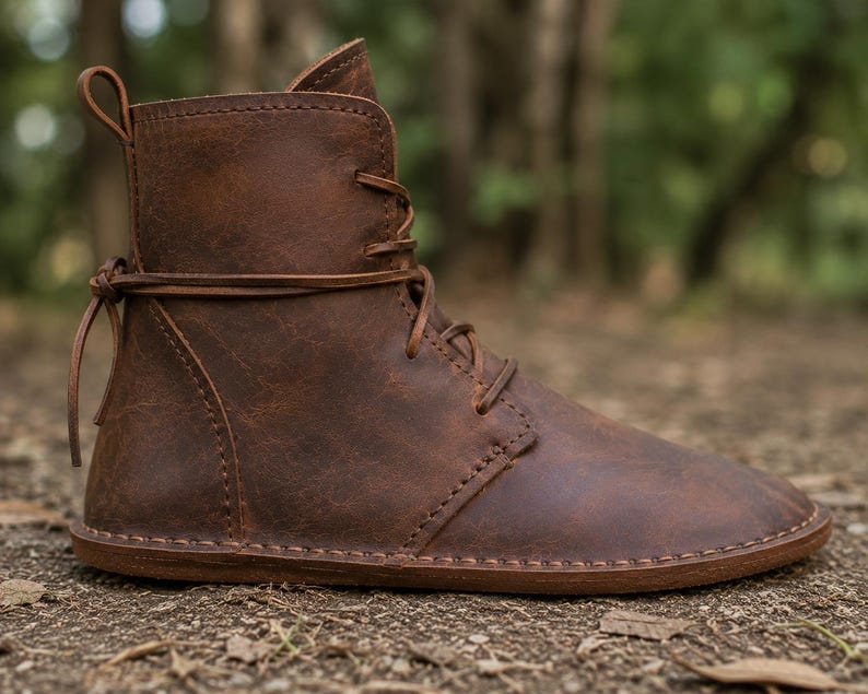 May include: A brown leather ankle boot featuring a rounded toe, a flat sole, and a lace-up closure that wraps around the ankle. The boot has visible stitching along the seams and a small pull tab at the top of the heel for ease of use.