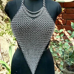 May include: A silver chainmail top displayed on a black mannequin. The top features a halter neck design with multiple chain layers and a triangular shape. The mannequin is set against a backdrop of brick and greenery.