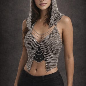 Puede incluir: Top de cota de malla plateada con capucha a juego. El top sin mangas tiene un escote pronunciado y se combina con pantalones negros. La cota de malla está hecha de pequeños anillos metálicos entrelazados. El fondo es gris oscuro.
