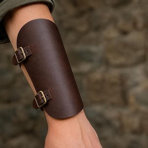 May include: A dark brown leather forearm bracer featuring two adjustable straps with antique brass-toned metal buckles. The smooth, curved piece of leather wraps around the forearm, secured by the buckles on the side for a snug fit.