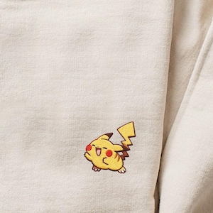 Puede incluir: Sudadera beige con un pequeño diseño bordado de Pikachu en la parte delantera. El Pikachu es amarillo con mejillas rojas y una cola en forma de rayo. La sudadera tiene cuello redondo y mangas largas.