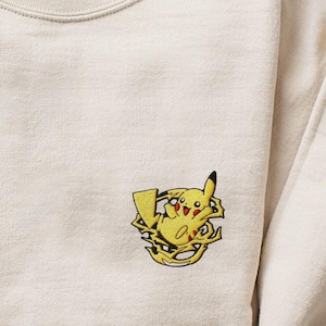 May include: Cream-colored sweatshirt with a crew neck and long sleeves. The sweatshirt features an embroidered Pikachu design in yellow, black, and red on the front. The fabric appears soft and comfortable.
