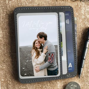 May include: A grey bi-fold wallet featuring a photograph of a couple embracing, with the text "I still fall for you every day." The wallet has card slots and a stitched border. A pen and a coin are visible.