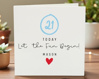 May include: A white greeting card with the number 21 in a blue circle, the words "TODAY Let the Fun Begin!" and the name "MASON" with a red heart. The card is on a wooden surface.
