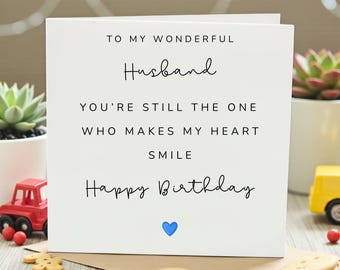 May include: A white greeting card with the text "TO MY WONDERFUL Husband YOU'RE STILL THE ONE WHO MAKES MY HEART SMILE Happy Birthday" in black script. A blue heart is at the bottom. The card is on a brown envelope.