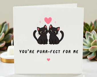 May include: A white greeting card with the text "YOU'RE PURR-FECT FOR ME" and two black cat illustrations. A pink heart and smaller pink hearts are also featured on the card.