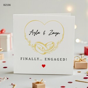 Könnte beinhalten: Eine quadratische weiße Grußkarte mit einem goldenen Herz-Umriss, der die Namen Ayla & Zayn über zwei ineinander verschlungenen goldenen Ringen umschließt. Darunter steht der Text FINALLY... ENGAGED! mit einem kleinen roten Herz-Symbol.