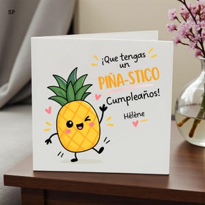 May include: A square white greeting card featuring a cartoon pineapple with a winking face, green leaves, and black limbs. The text reads ¡Que tengas un PIÑA-STICO Cumpleaños! Hélène, accented with small pink hearts and yellow dash marks.