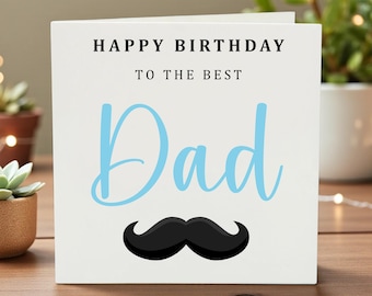 May include: A white greeting card with the text "HAPPY BIRTHDAY TO THE BEST Dad" in black and light blue. A black mustache graphic is below the word "Dad". The card is on a wooden surface.