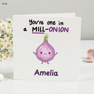 May include: A square white card featuring the phrase "You're one in a MILL-ONION" in black and purple. A cartoon onion with a smiling face is below the text. The name "Amelia" is written in purple at the bottom.