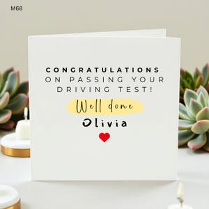 May include: A white greeting card with the text "CONGRATULATIONS ON PASSING YOUR DRIVING TEST!" in black. Below, "Well done" is written in a script font over a yellow brushstroke, with "Olivia" and a red heart below.