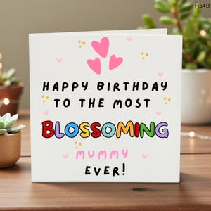 May include: A white birthday card with the text "HAPPY BIRTHDAY TO THE MOST BLOSSOMING mummy EVER!" in colorful letters. The card features pink hearts and yellow star accents. The card is on a wooden surface.