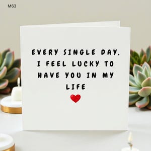 Könnte beinhalten: Weiße Grußkarte mit schwarzem Text: "EVERY SINGLE DAY, I FEEL LUCKY TO HAVE YOU IN MY LIFE". Unten befindet sich ein kleines rotes Herz. Die Karte ist von Sukkulenten und Kerzen umgeben.