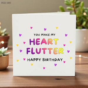 May include: A white birthday card with the message "You Make My Heart Flutter Happy Birthday" in colourful letters. Small purple, yellow, and pink hearts are scattered around the text. The card is on a wooden surface.