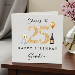 以下が含まれることがあります： 黒い筆記体のCheers To、金色のグリッターで描かれた大きな数字の25、金色の風船、シャンパンボトル、2つのシャンパングラス、黒い文字のHAPPY BIRTHDAY、そして黒い筆記体のSophiaという名前が印刷された白い正方形のグリーティングカード。