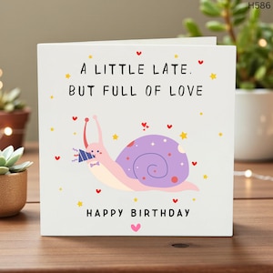 May include: A white birthday card with the text "A LITTLE LATE. BUT FULL OF LOVE" and "HAPPY BIRTHDAY". The card features a cartoon snail with a purple shell, holding a party hat, surrounded by hearts and stars.