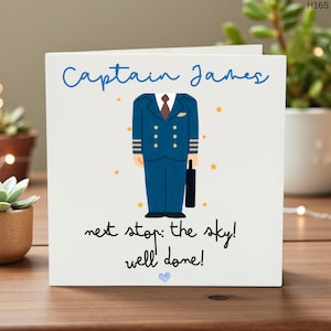 May include: A white card with the words "Captain James" in blue script. It features a pilot's uniform illustration and the phrases "next stop: the sky! well done!" and a blue heart. The card is on a wooden surface.