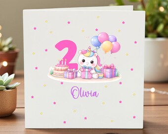May include: A white birthday card featuring a colorful illustration of a unicorn holding a gift, with a number 2, balloons, and a cake. The name "Olivia" is written in purple script. Pink and yellow stars are scattered around the card.