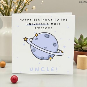 May include: A white birthday card with the text "HAPPY BIRTHDAY TO THE UNIVERSE'S MOST AWESOME UNCLE!" features a light blue planet with rings and star accents. The card is decorated with small blue stars.