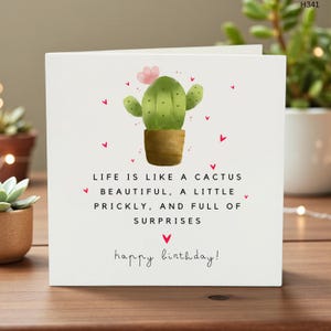 May include: A white birthday card featuring a green cactus in a brown pot, pink flower, and red hearts. The card has the text: "Life is like a cactus, beautiful, a little prickly, and full of surprises. Happy birthday!"