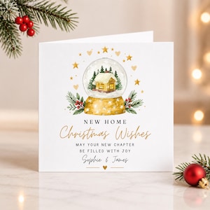 Puede incluir: Una tarjeta de felicitación cuadrada blanca con una bola de nieve en acuarela que contiene una casa y pinos. El texto dice NEW HOME Christmas Wishes MAY YOUR NEW CHAPTER BE FILLED WITH JOY Sophie & James. Estrellas y corazones dorados rodean la bola.