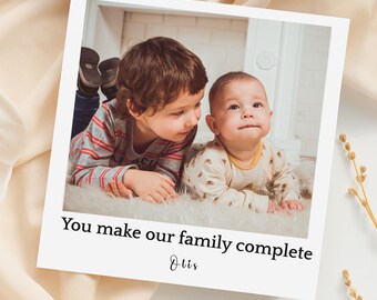 May include: A white polaroid-style photo featuring two children. One child is wearing a striped shirt, and the other is in a patterned onesie. The photo has the text "You make our family complete" and the name "Otis".