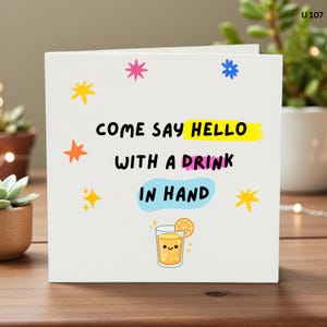 May include: A white greeting card with the text "COME SAY HELLO WITH A DRINK IN HAND" in black, yellow, and pink. The card features colorful star and flower illustrations and a cartoon glass of orange juice with a smiling face and a lemon slice.