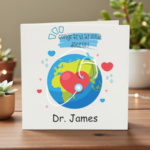 Può includere: Biglietto di auguri bianco con un design colorato. La carta presenta un globo con un cuore rosso, uno stetoscopio e le parole "congratulations doctor!" in blu. Il nome "Dr. James" è stampato in basso.