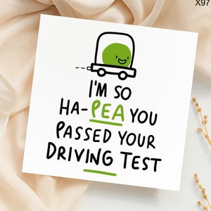 May include: A white greeting card with the text "I'M SO HA-PEA YOU PASSED YOUR DRIVING TEST" in black and green. The card features a cartoon pea driving a car. The background is a light beige fabric.