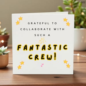 May include: A white greeting card with the text "Grateful to collaborate with such a fantastic crew!" in black and yellow lettering. The card is decorated with yellow star illustrations and small pink hearts.