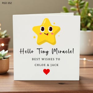 Pode incluir: Um cartão branco com o texto "Hello Tiny Miracle!" e "Best Wishes to Chloe & Jack." Uma estrela amarela de desenho animado com um rosto sorridente está no topo, e um coração vermelho na parte inferior. O cartão está em uma superfície de madeira.