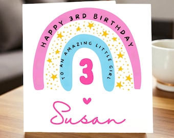 Op de afbeelding: Een witte verjaardagskaart met een roze en blauw regenboogontwerp. De kaart leest "Happy 3rd Birthday To An Amazing Little Girl" met een groot roze nummer 3. De naam "Susan" staat eronder in roze script.
