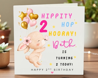 May include: A square birthday card with a cartoon bunny holding balloons. The card reads "Hippity Hop Hooray! Beth is turning 2 today! Happy 2nd Birthday" in pink, yellow, and black text.