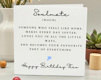 May include: A white greeting card with the word "Soulmate" and a definition. The card includes the text "Happy Birthday Theo" and a blue heart. A small succulent plant and wooden object are visible in the background.