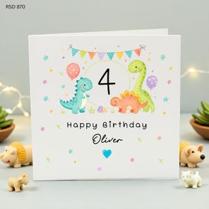 May include: White birthday card featuring a colorful dinosaur theme. The card displays the number 4, the words "Happy Birthday Oliver", and illustrations of dinosaurs, balloons, and stars. A blue heart is also present.