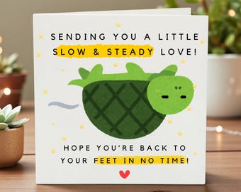 May include: A white greeting card with a cartoon turtle illustration. The card has text that reads "SENDING YOU A LITTLE SLOW & STEADY LOVE! HOPE YOU'RE BACK TO YOUR FEET IN NO TIME!" with a red heart at the bottom.