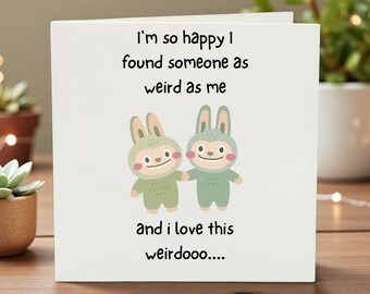 May include: A white greeting card with the text "I'm so happy I found someone as weird as me and I love this weirdooo...." Two cartoon bunnies in green outfits are holding hands. The card is on a wooden surface.