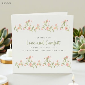 Può includere: Un biglietto di auguri bianco con decorazioni floreali e la scritta "Sending You Love and Comfort". Il biglietto ha un bordo floreale ed è circondato da piante grasse e candele.