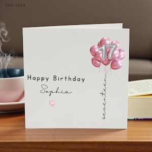 May include: A white square greeting card featuring the text Happy Birthday Sophia with a small pink heart. A cluster of pink balloons is tied to a string with the word seventeen written vertically, and the silver number 17 is centred.