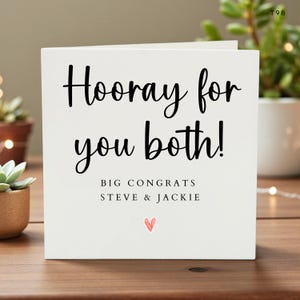 May include: A white greeting card with the words "Hooray for you both!" in black script. Below, it reads "BIG CONGRATS STEVE & JACKIE" with a small red heart. The card is on a wooden surface with plants in the background.