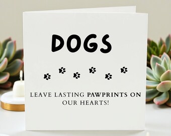 May include: A white greeting card with the word "DOGS" in large, black letters. Below the word are six black paw prints. The text "LEAVE LASTING PAWPRINTS ON OUR HEARTS!" is printed below the paw prints.
