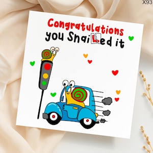 May include: A white greeting card with a cartoon illustration. The image features a snail driving a blue car, a traffic light, and the text "Congratulations you Shailed it" in red and black. Colorful hearts are scattered around.