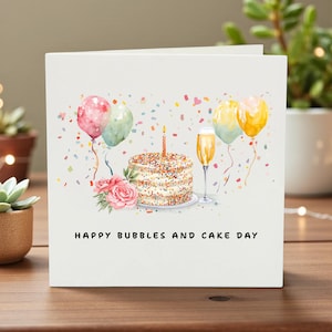 May include: A square greeting card featuring a watercolour illustration of a birthday cake with a single candle, a glass of champagne, colourful balloons, and pink roses. The text HAPPY BUBBLES AND CAKE DAY is printed in black at the bottom.