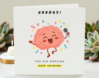 May include: A white greeting card with a cartoon brain holding a microphone and confetti. The card reads "HOORAY!" at the top, with "YOU DID AMAZING" and "KEEP SHINING" at the bottom. The brain is pink with a smiling face.
