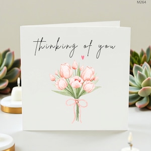 Può includere: Biglietto di auguri bianco con la scritta "thinking of you" in corsivo nero. Il biglietto presenta un'illustrazione ad acquerello di un bouquet di tulipani rosa legato con un nastro rosa. Un piccolo cuore rosa è sopra il bouquet.