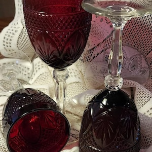 May include: Ruby red wine glasses with intricate cut glass patterns. The glasses have a diamond and leaf design, with a clear stem and base. One glass is upright, while another is lying on its side. The glasses are on a white lace tablecloth.