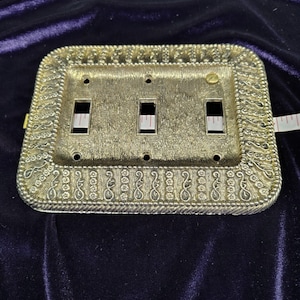 May include: A rectangular gold-toned metal triple light switch wall plate with a textured surface and a decorative border featuring floral and scroll patterns. A white measuring tape is visible through the three rectangular switch openings.