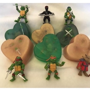 May include: A selection of Teenage Mutant Ninja Turtles action figures and a Shredder figure, arranged on and around heart-shaped objects. The figures are green, red, and brown, with weapons. The hearts are in shades of green and tan.